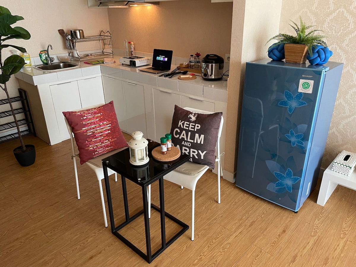 A compact kitchen area is visible, featuring a sink and various kitchen essentials. A small black table is surrounded by two chairs, adorned with decorative pillows. Nearby, a blue refrigerator adds a pop of color to the arrangement, with a plant accentuating the space.