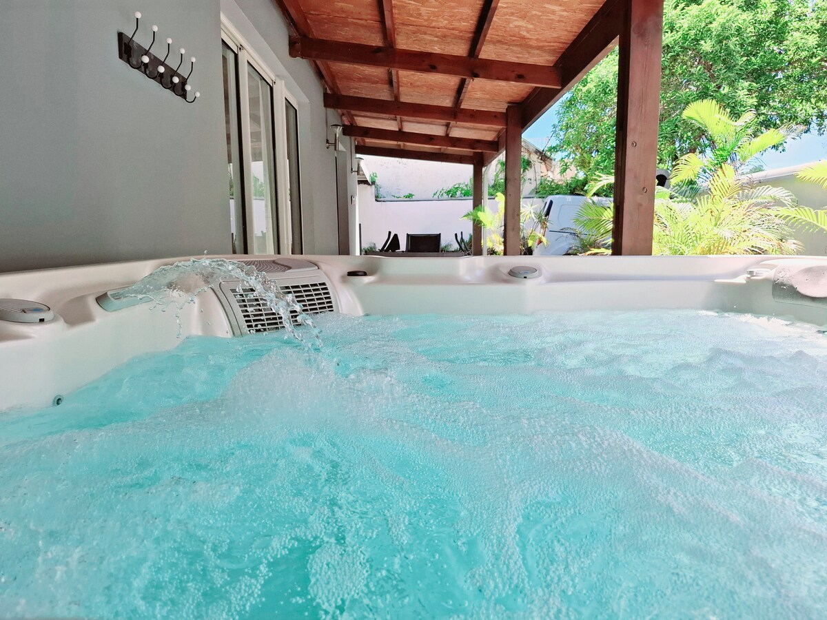 A relaxing jacuzzi is positioned under a wooden pergola, with bubbles rising in the inviting water. The structure is flanked by greenery, and the interior of the home is visible through glass doors, merging indoor comfort with outdoor tranquility.