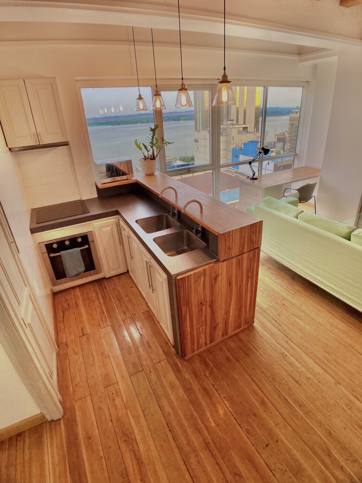 The kitchen features minimalist wooden cabinets and a countertop with two sinks. Large windows provide views of the city and river, allowing natural light to fill the space. Pendant lights hang above, enhancing the contemporary feel of the area.