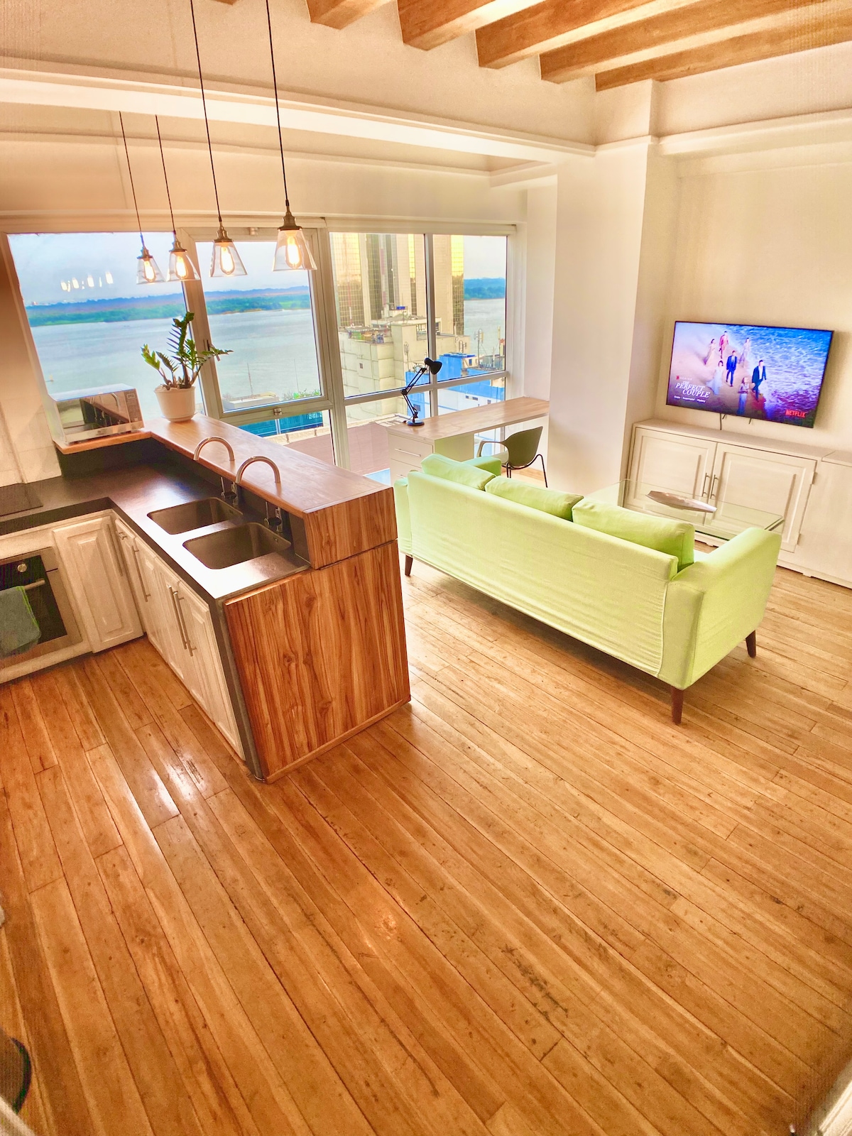 An open living area showcases a light green sofa facing a flat-screen TV, with large windows providing views of the city and river. The kitchen features wooden cabinetry and a modern sink, complemented by polished hardwood teak floors that reflect natural light.