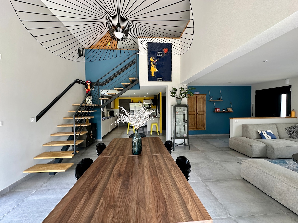 An open living area is depicted with a large wooden dining table set at the forefront. A minimalist staircase with wooden steps leads to an upper level. The background features a modern kitchen and lounge area, with stylish decor elements including artwork and plants.