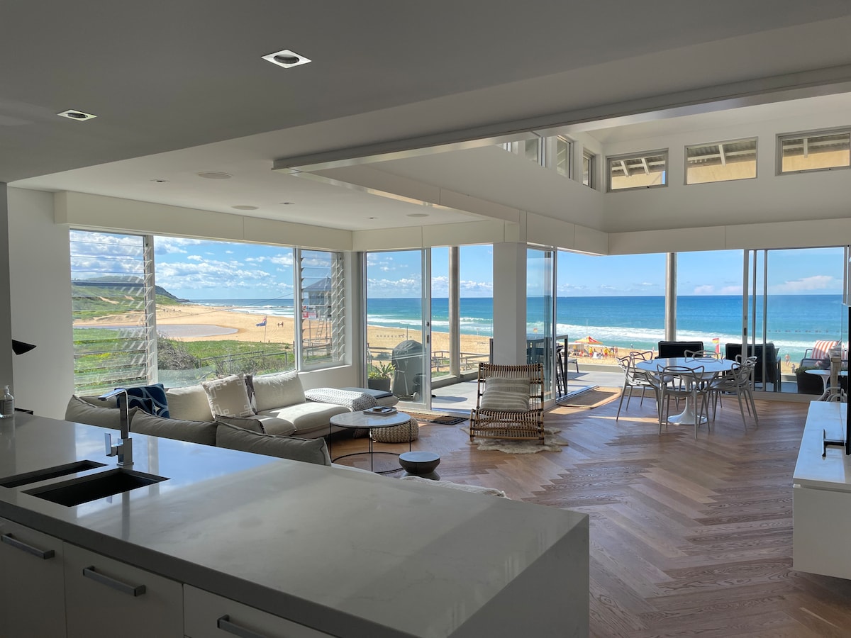 An open-plan living area is highlighted by large windows offering expansive views of the beach and ocean. A spacious light-filled lounge features comfortable seating and a dining area, with natural wood flooring contributing to the airy feel. Fresh coastal scenery complements the modern design.