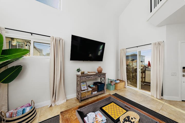 A Sleek Smart TV is Mounted above a Media Console. Natural Textures & Sheer Curtains Enhance the Airy Vibe, while Nautical decor and Soft Lighting Complete the Relaxed, Seaside Atmosphere