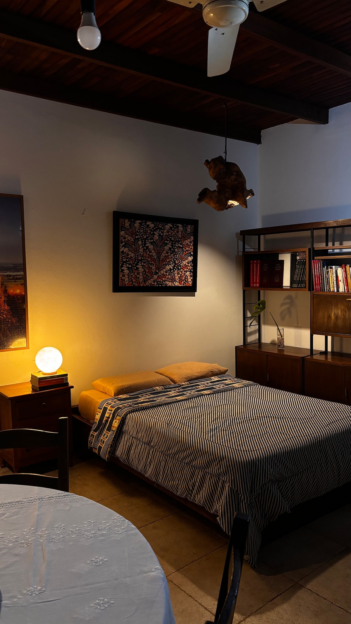 A cozy sleeping area features a comfortably made bed with striped bedding, complemented by soft lighting from a spherical lamp. A wooden bookshelf, arranged with books, stands nearby, and a piece of wall art adds a touch of character to the room.