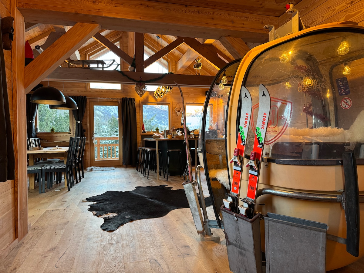 An open-plan living area features wooden beams and a large window offering views of the mountains. A ski gondola is positioned next to rustic furnishings, including a dining table and black chairs. A cowhide rug is placed on the wooden floor, enhancing the chalet's cozy ambiance.