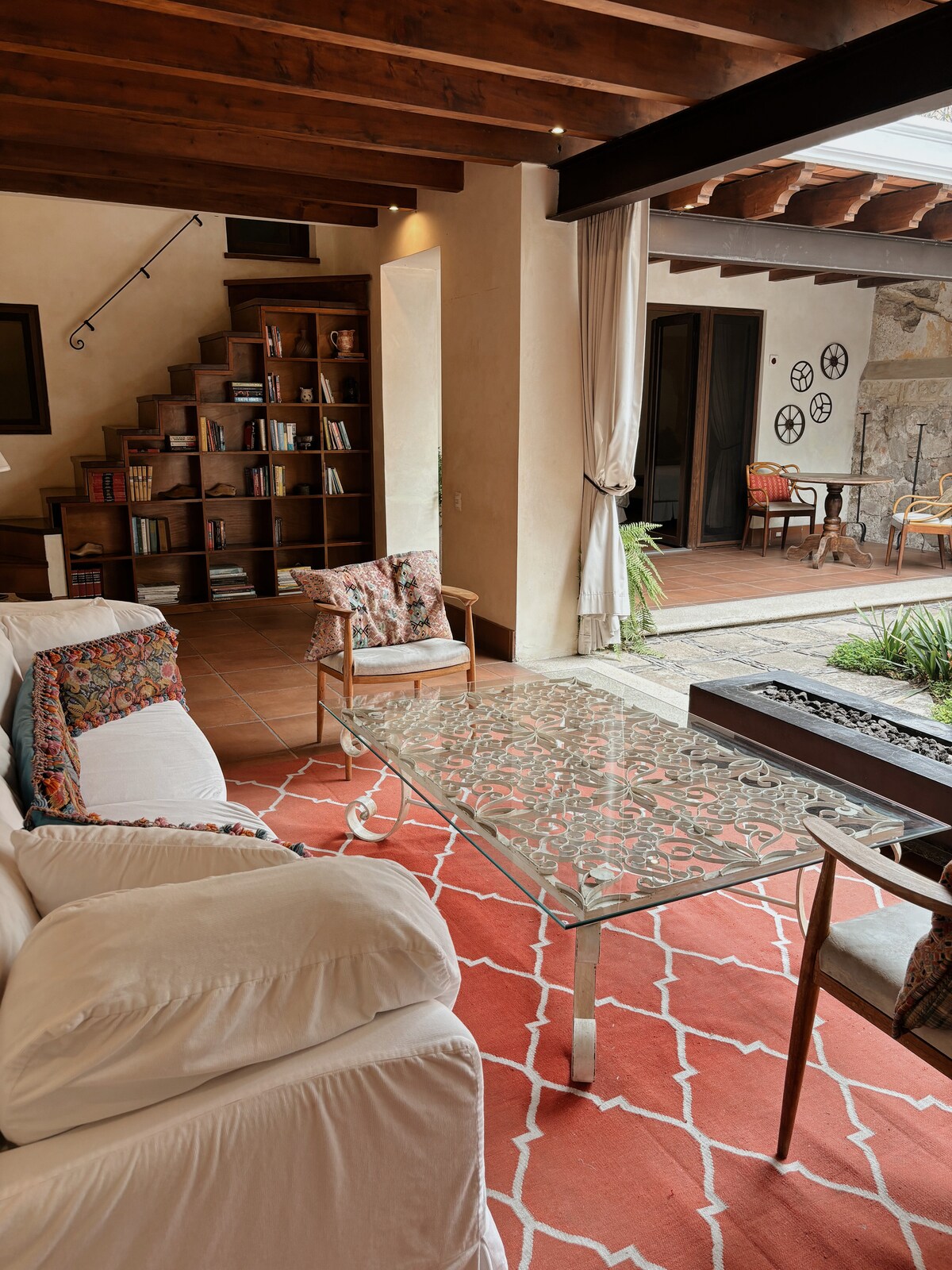 An inviting living space features a light-colored sofa with colorful throw pillows, paired with two wooden chairs. A glass coffee table rests on a patterned rug, while an open staircase leads to a bookshelf. Natural light streams through large glass doors, enhancing the airy ambiance.