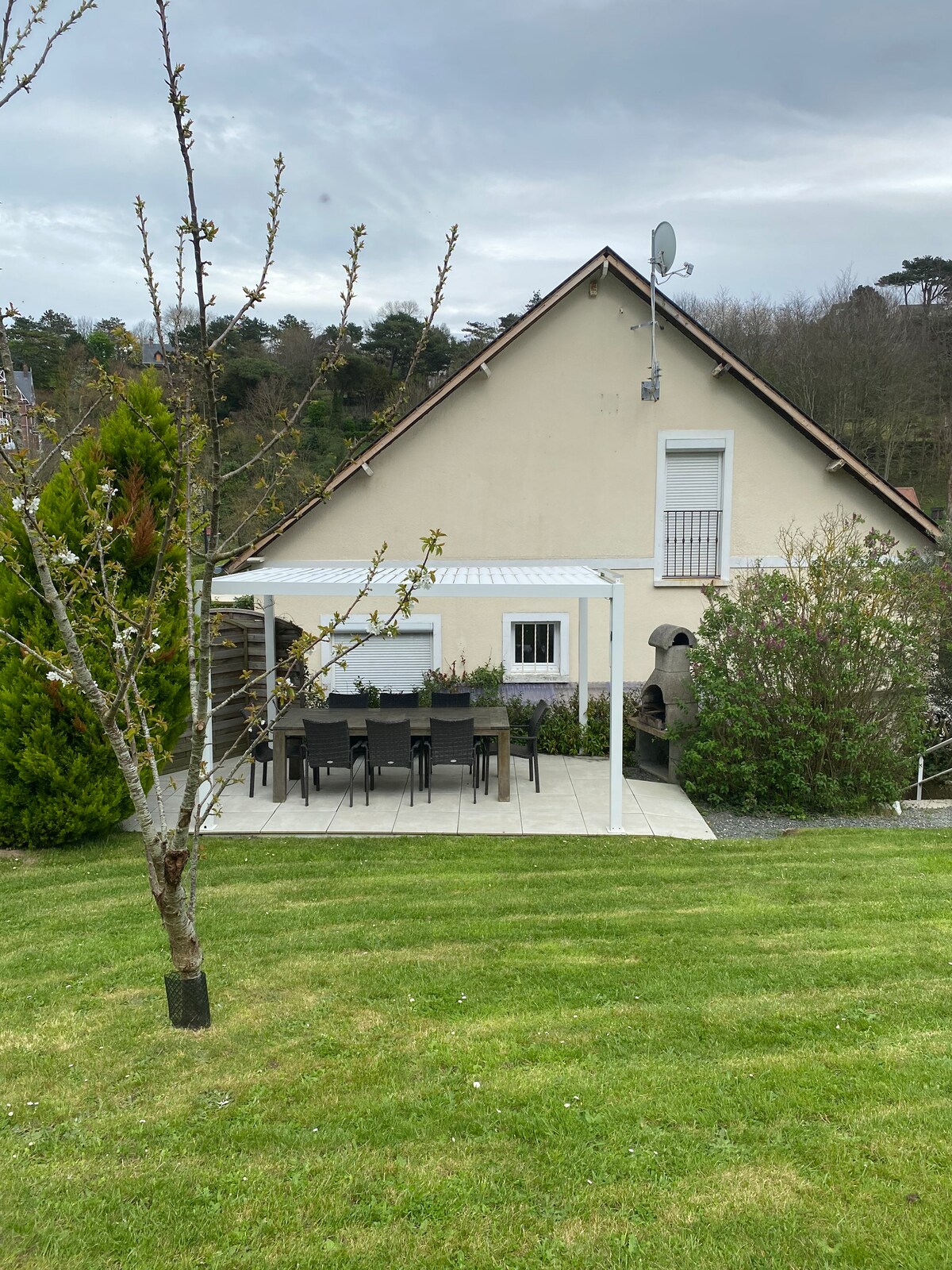 An outdoor terrace area is visible, featuring a dining set with several chairs positioned beneath a covered structure. The well-maintained lawn is framed by a variety of shrubs and small trees, while a neutral-colored house is situated in the background.