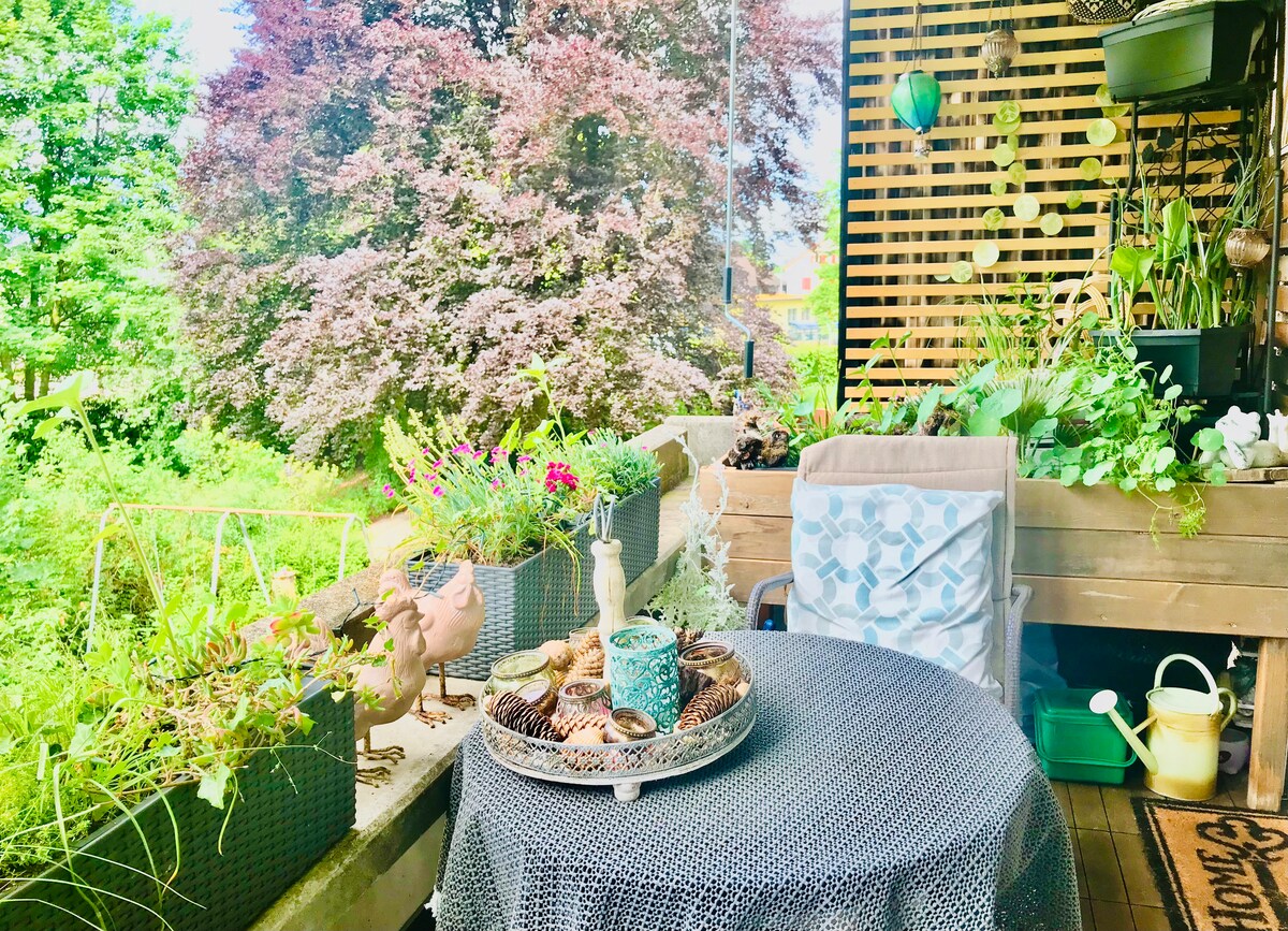 A balcony adorned with a round table and two comfortable chairs is surrounded by a variety of potted plants and flowers. Natural light highlights the greenery, and decorative items, including a tray with pinecones, add a touch of charm.
