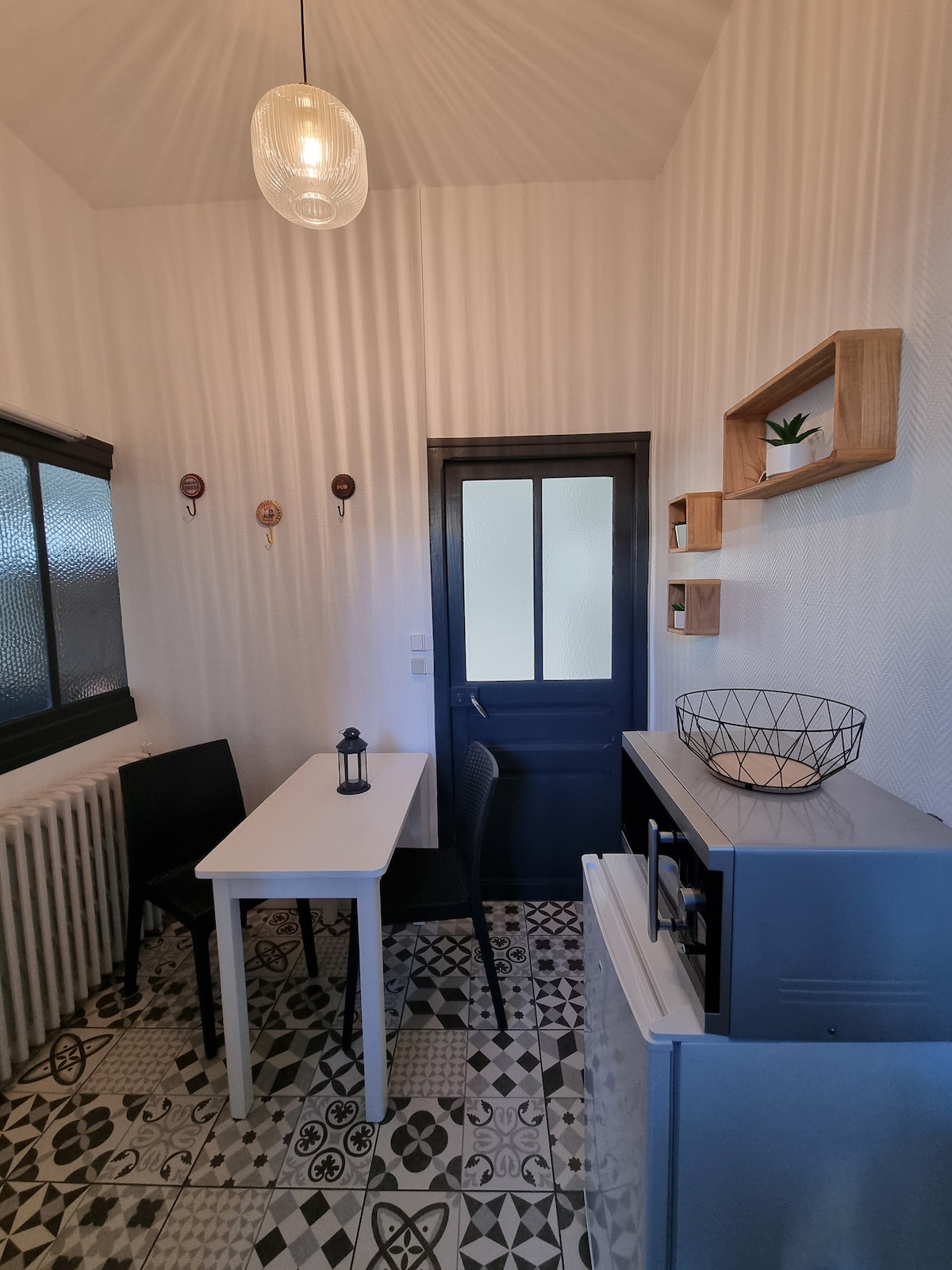 A compact kitchen area is highlighted by patterned tile flooring and textured walls. A rectangular table is surrounded by black chairs, and a small decorative shelf with a plant is mounted on the wall. A glass door and a fridge enhance the functional layout of the space.