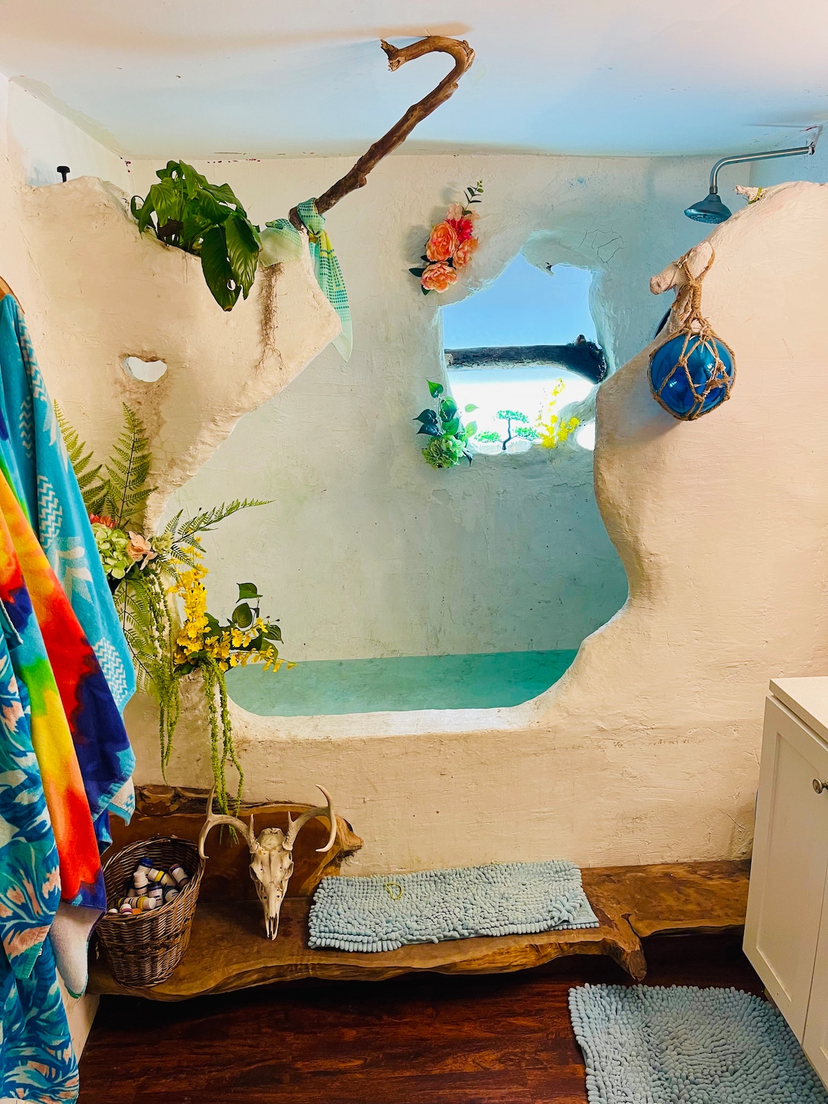 The bathroom features an artistic grotto-style bathtub with unique organic shapes and a soothing turquoise water surface. Natural light filters through a window adorned with tropical plants. Decorative elements include hanging foliage, a skull, and a woven basket, enhancing the space's creative design.