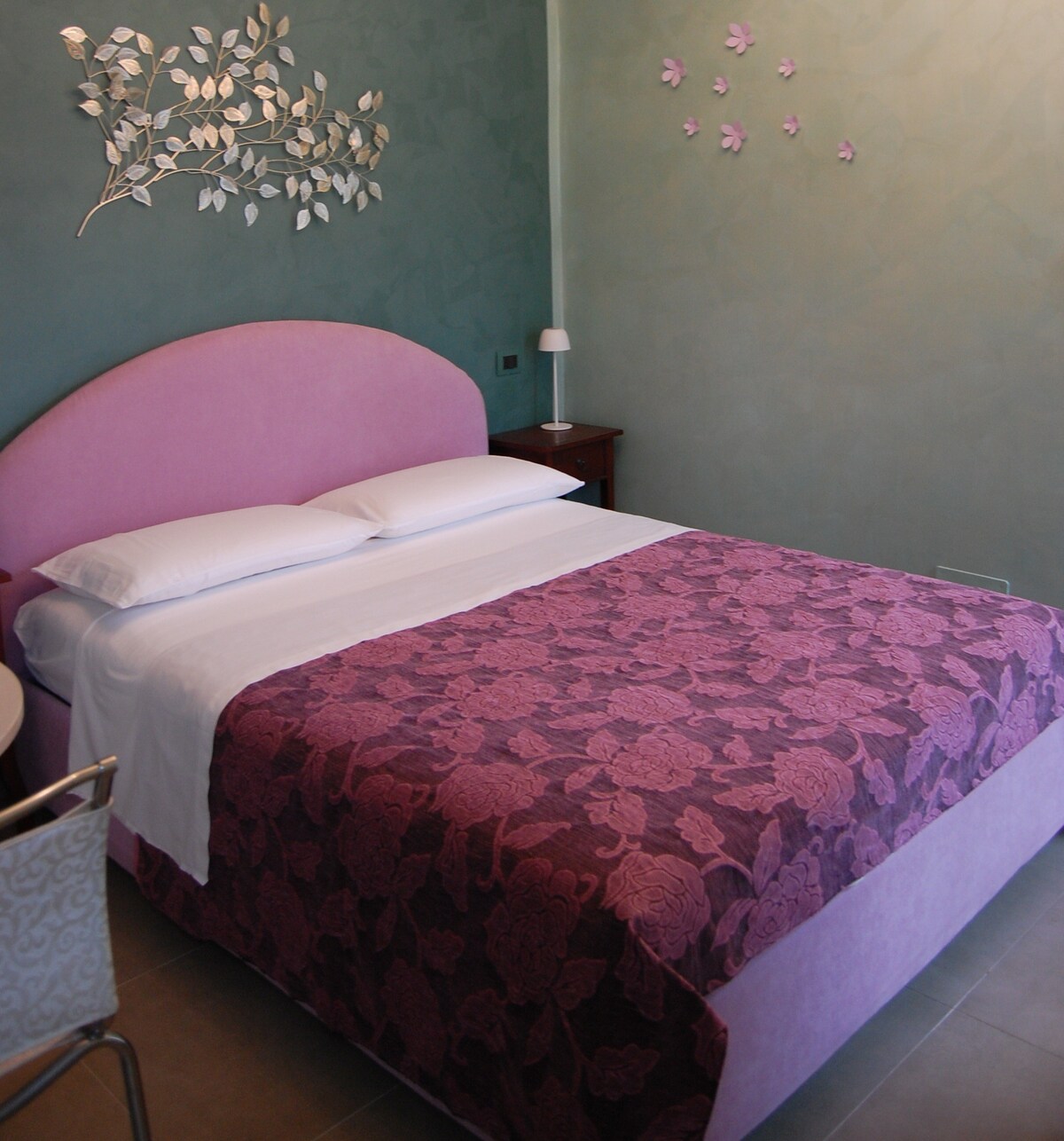 A sleeping area features a spacious bed dressed in elegant bedding with floral patterns. The walls are accented in soft green, complemented by decorative elements and subtle wall art. A small bedside table is positioned next to the bed, offering a minimalist design.