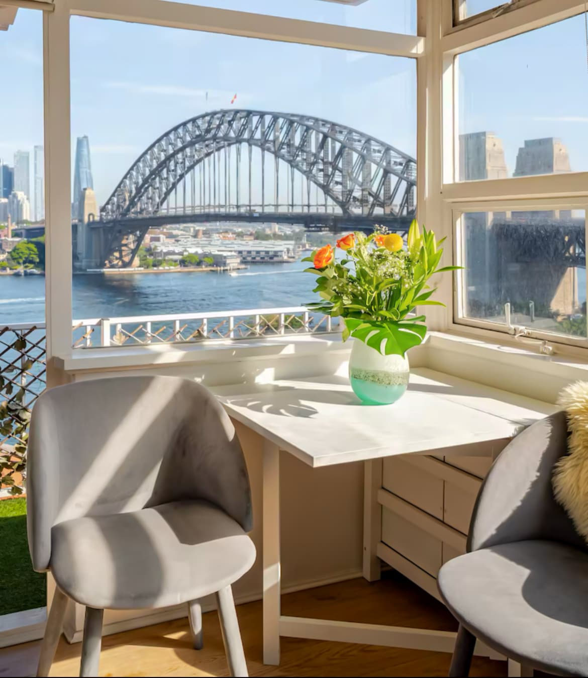 A bright corner dining nook features a small white table paired with two grey chairs. A vase of colorful flowers sits on the table, complementing the stunning view of the Sydney Harbour Bridge visible through large windows.