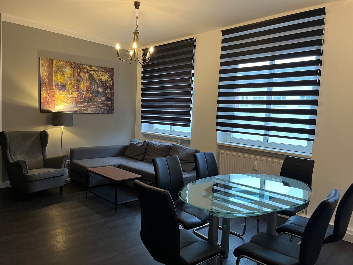 A modern living space is showcased with large windows covered by striped roller shades, allowing for natural light. A contemporary glass dining table is surrounded by black chairs. A gray sofa and an armchair are positioned near a wall adorned with a framed art piece.