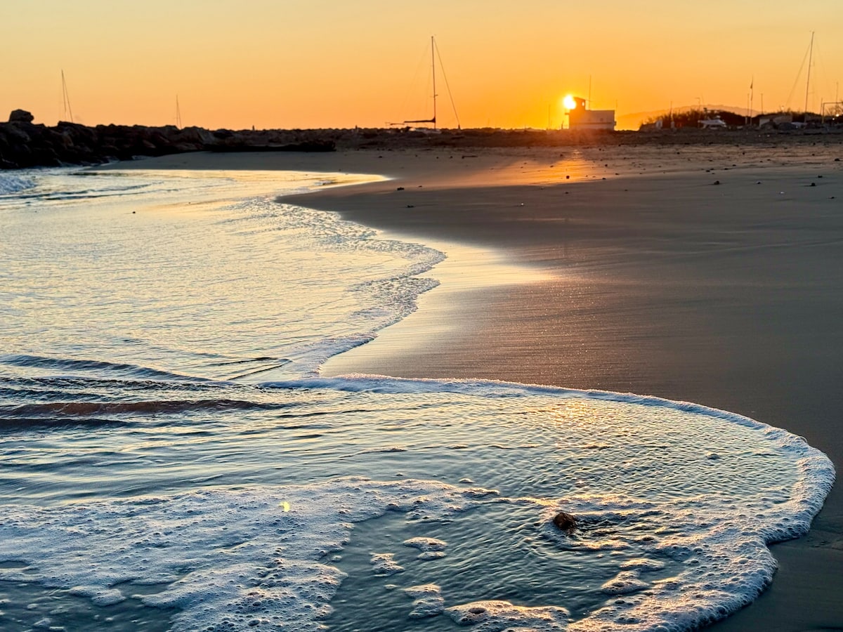 The image captures a serene beach scene at sunset, with gentle waves lapping at the shore. The warm colors of the sky reflect on the water’s surface, alongside distant silhouettes of boats anchored in the harbor.
