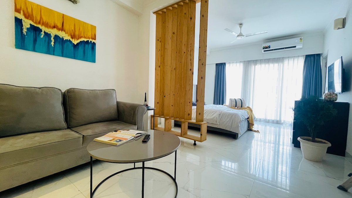 A spacious living area features a grey sofa and a modern circular coffee table. A wooden partition separates the sleeping area, where a super king bed is situated. Bright natural light filters through large glass doors that lead to a balcony, enhancing the airy ambiance.