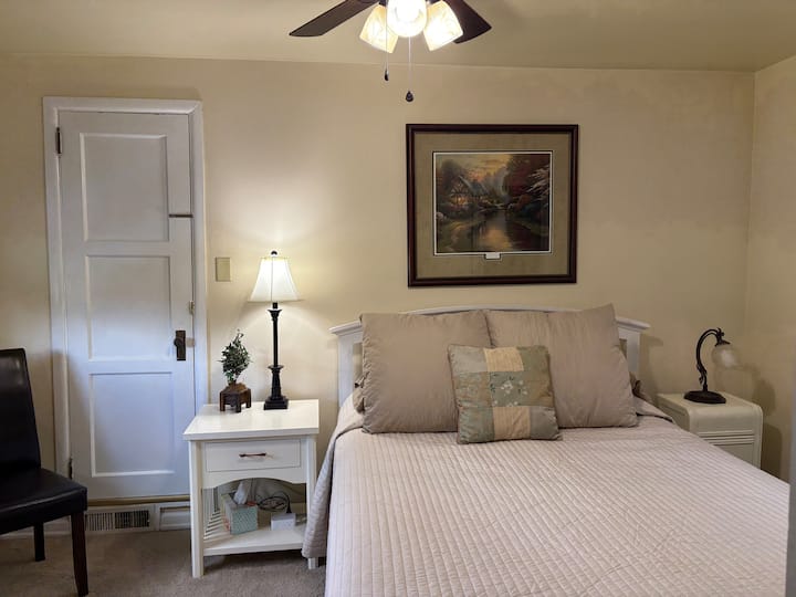 Dormitorio con cama tamaño queen, mesita de noche, silla cómoda, cómoda, calefacción por zócalo y enfriador de ventanas.