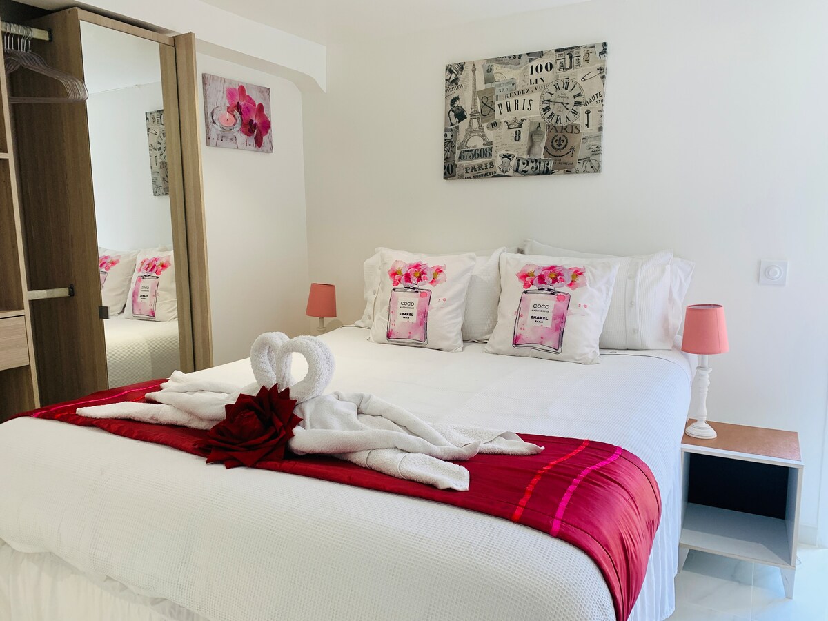 A spacious sleeping area features a large bed adorned with decorative pillows and a cozy blanket at the foot. A wooden nightstand holds a pink lamp, and a piece of wall art adds character. Reflections from a mirrored wardrobe create an airy feel.