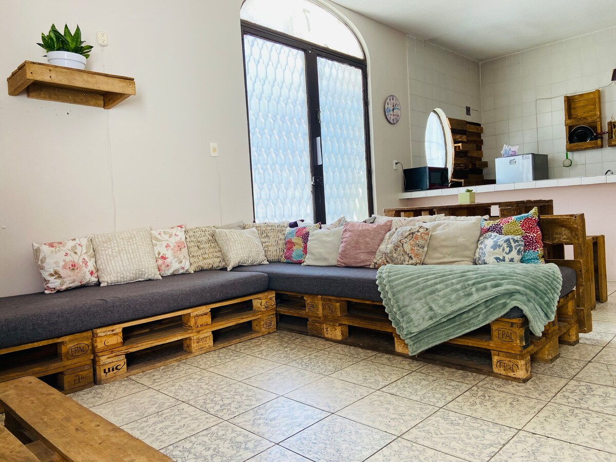 A spacious seating area is created with a large sofa made from wooden pallets, topped with an assortment of decorative cushions and a soft, textured blanket. Natural light enters through a sizable window, illuminating the tiled floor and accentuating the welcoming atmosphere.