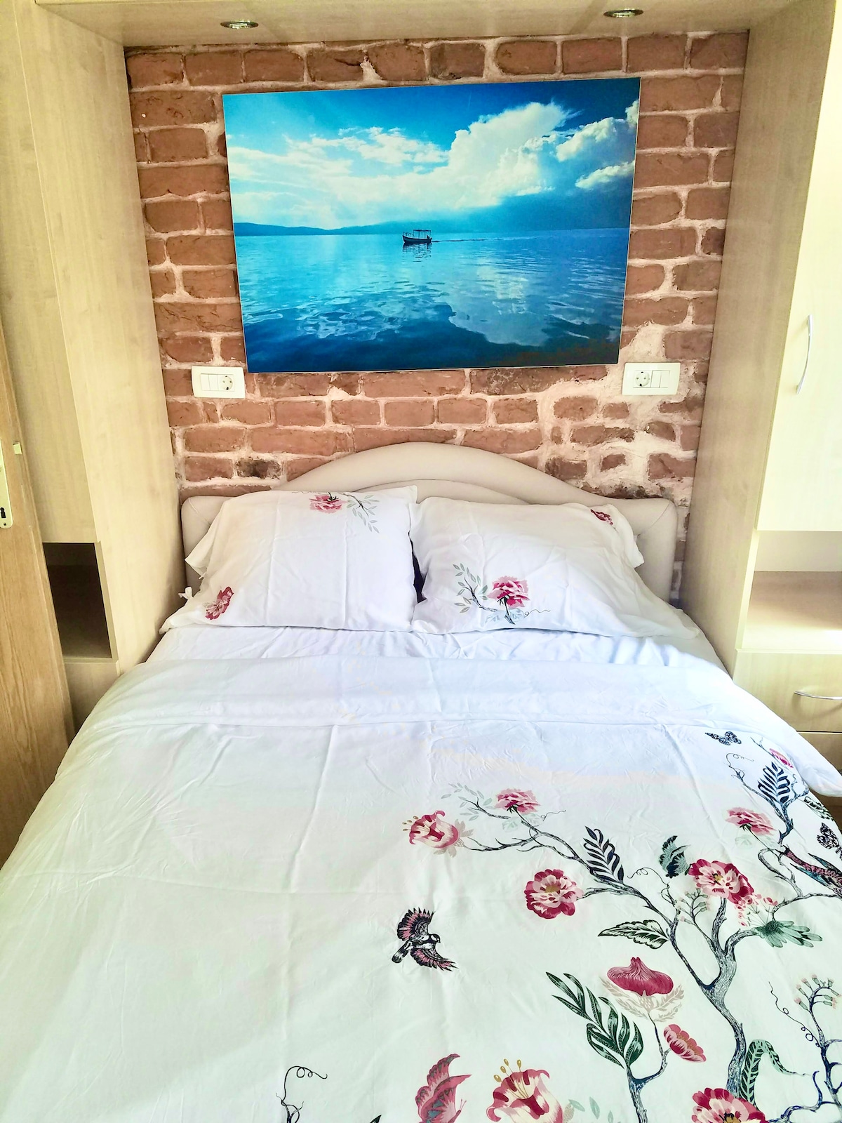 The sleeping area features a spacious bed dressed in light linens with floral patterns. A large artwork depicting a serene water scene and sky adds a calming element to the exposed brick wall backdrop, enhancing the overall aesthetic of the room.