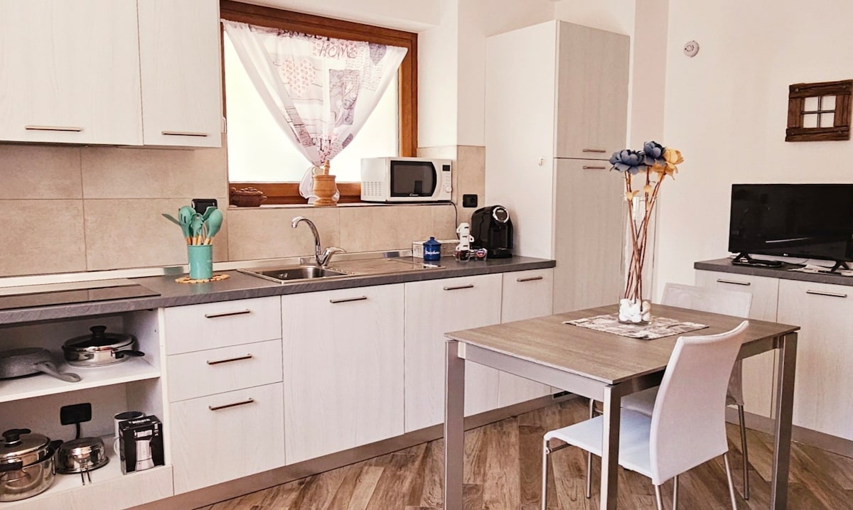 The kitchen space is equipped with modern appliances, including a microwave and coffee maker. A small dining table is set for two, surrounded by light-colored cabinetry. Decorative elements, such as a floral arrangement, add a touch of warmth to the functional layout.