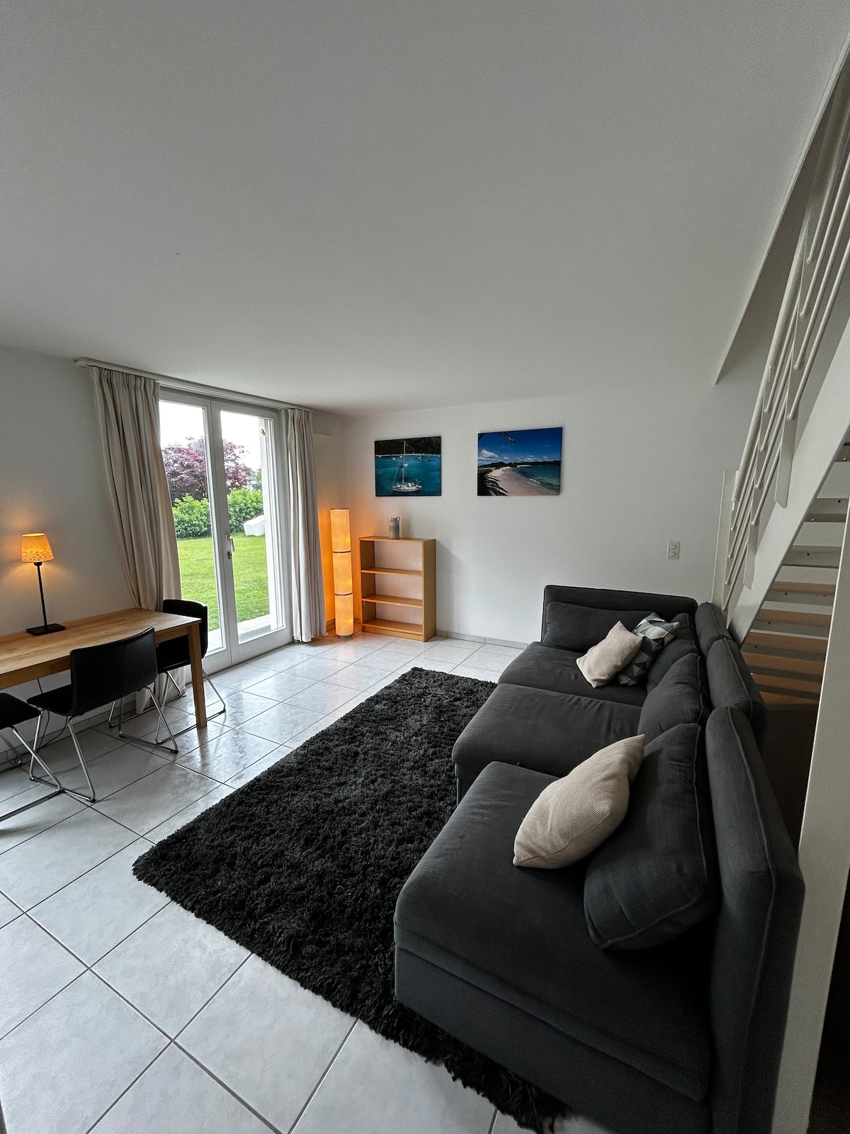 A cozy living area features a comfortable sofa with decorative cushions, a stylish table for dining or working, and a soft area rug. Natural light enters through large doors, overlooking the garden. A floor lamp and bookshelves complement the serene ambiance.