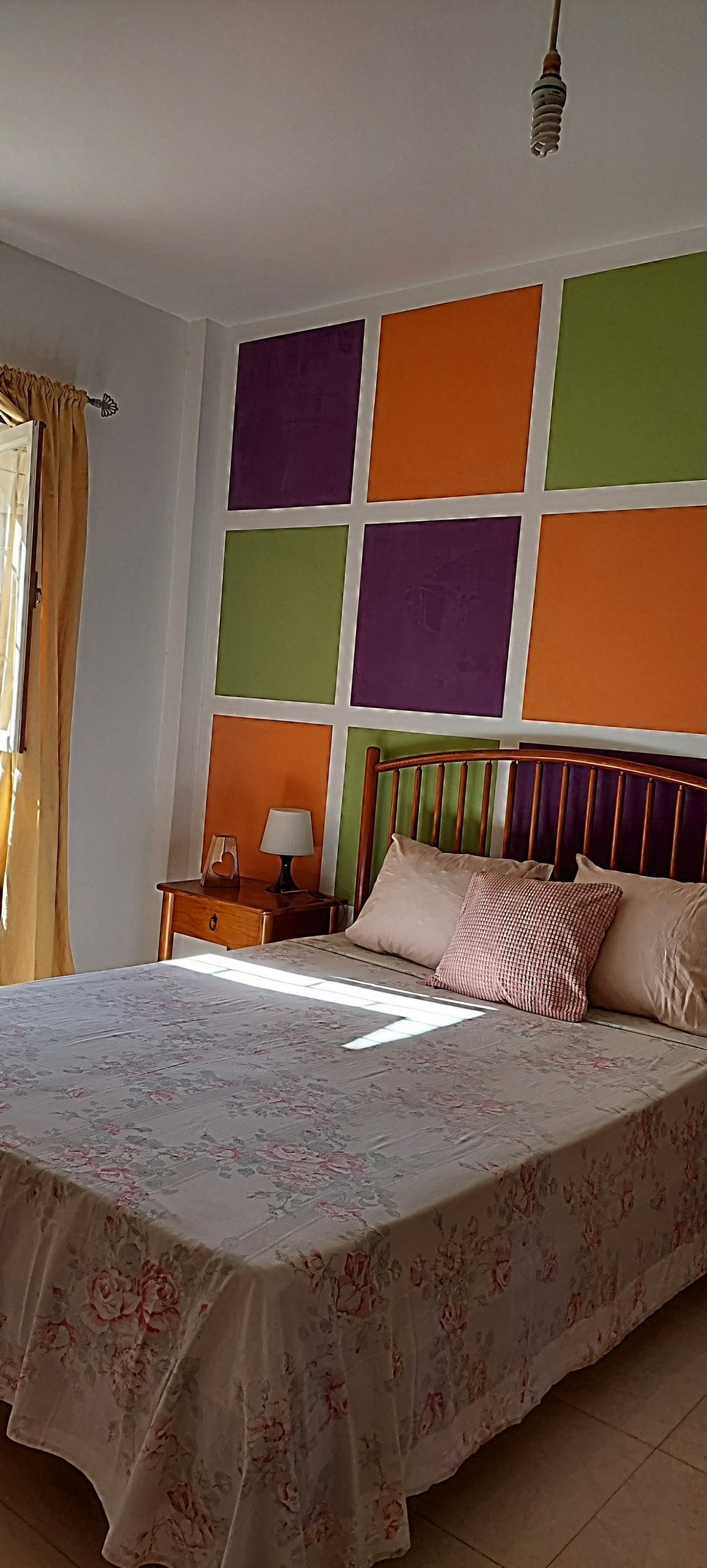 A bright bedroom features a spacious bed covered with a floral blanket. Colorful accent walls in orange, green, and purple add vibrancy. Sunlight streams through a window with light curtains, illuminating a bedside table with a lamp.