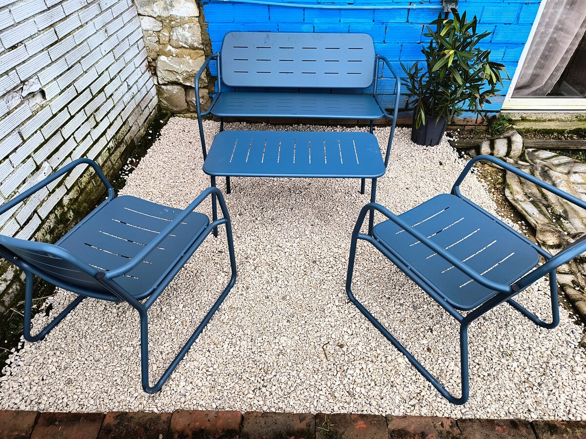 A seating area is presented with a blue metal sofa and two matching chairs, arranged around a central table. The gravel surface is complemented by a potted plant, with a textured wall painted in blue serving as a backdrop.