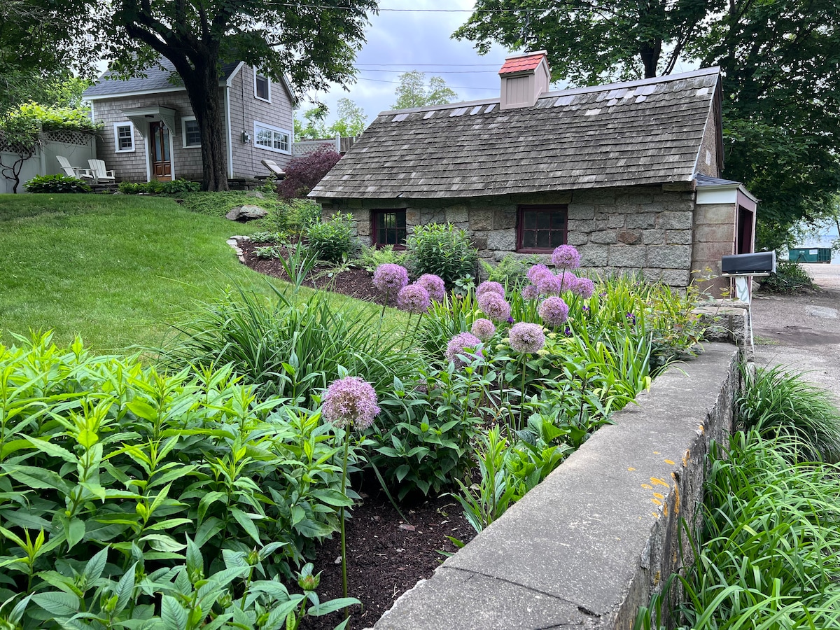 A quaint stone cottage is surrounded by lush greenery and colorful flowers, with a well-maintained garden featuring clusters of purple blooms. The cottage's rustic charm is complemented by a sloped lawn and mature trees, creating a serene outdoor atmosphere.