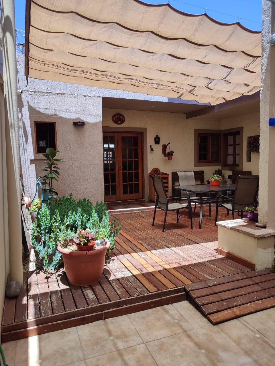 A cozy outdoor patio is bordered by wooden decking, complemented by potted plants and greenery. A shaded seating area with a table and chairs provides a comfortable space for relaxation and gathering, while a warm light filters through the open doors leading inside.