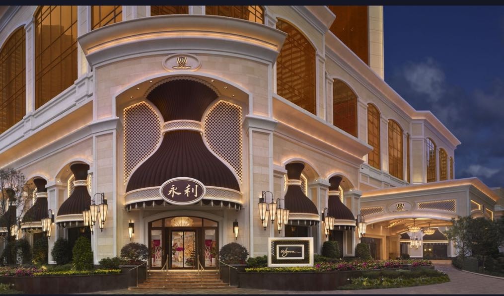 An elegantly designed entrance features a grand awning with warm lighting. Tall windows reflect the evening sky, while manicured plants adorn the pathway leading to the entrance.