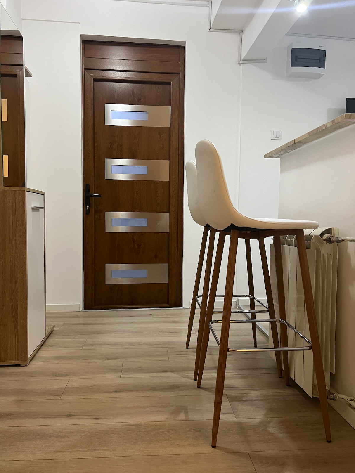 An interior view features a modern brown door with horizontal glass panels, leading to another space. Two high-backed white stools are positioned at a countertop, with light brown wood flooring extending throughout the room and a glimpse of cabinetry on the left.