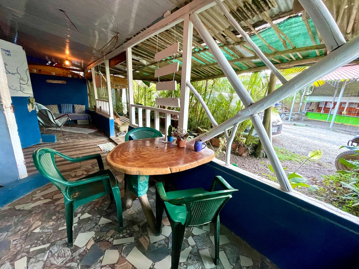 An outdoor dining area features a round wooden table surrounded by green plastic chairs. The space is sheltered by a partially open roof, allowing natural light to filter in. Lush greenery can be seen in the background, creating a relaxed atmosphere.