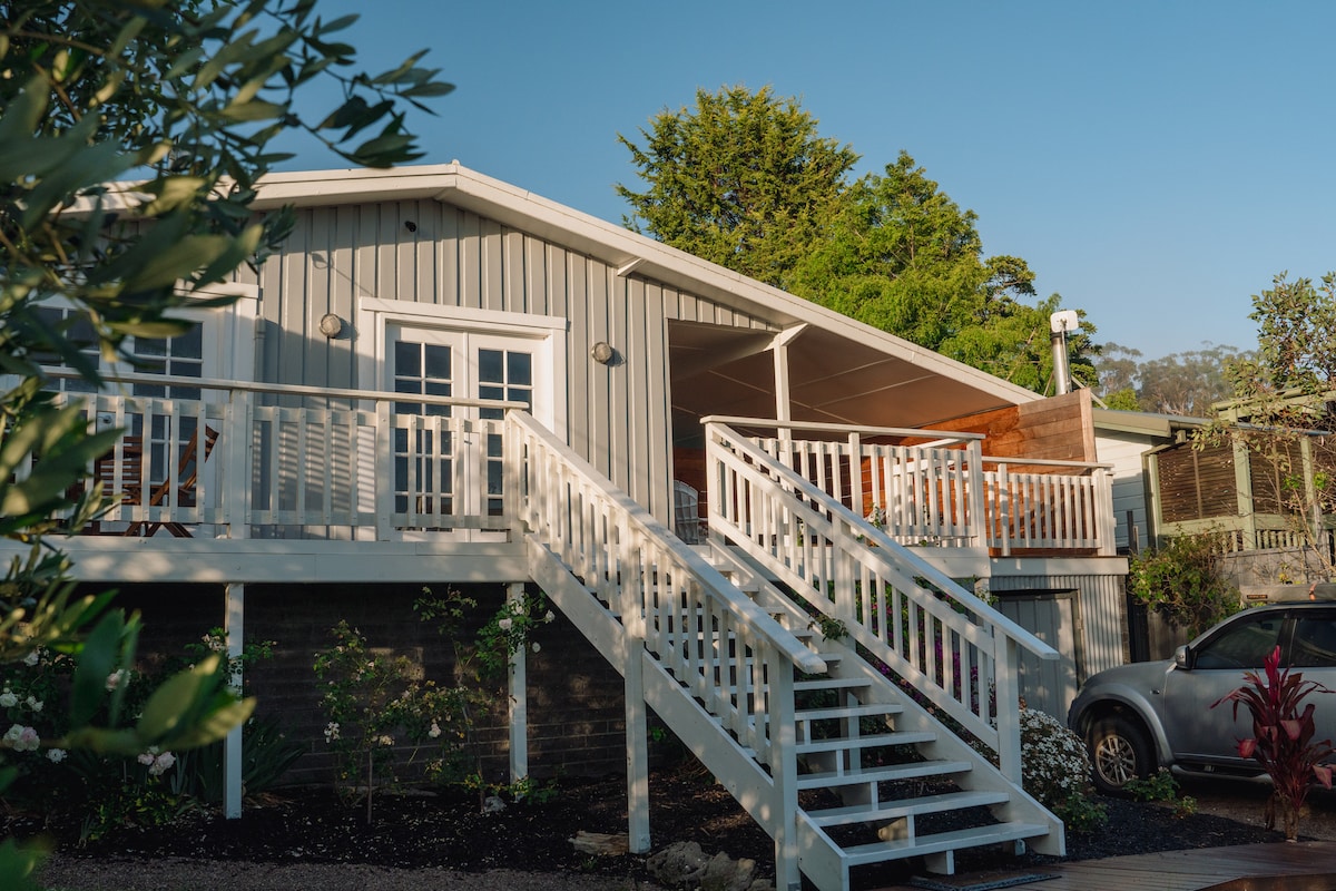The inviting exterior of a spacious beach house is highlighted by white railings and stairs leading to a covered porch. Surrounding greenery complements the neutral tones of the home, providing a tranquil entrance that blends with the natural landscape.