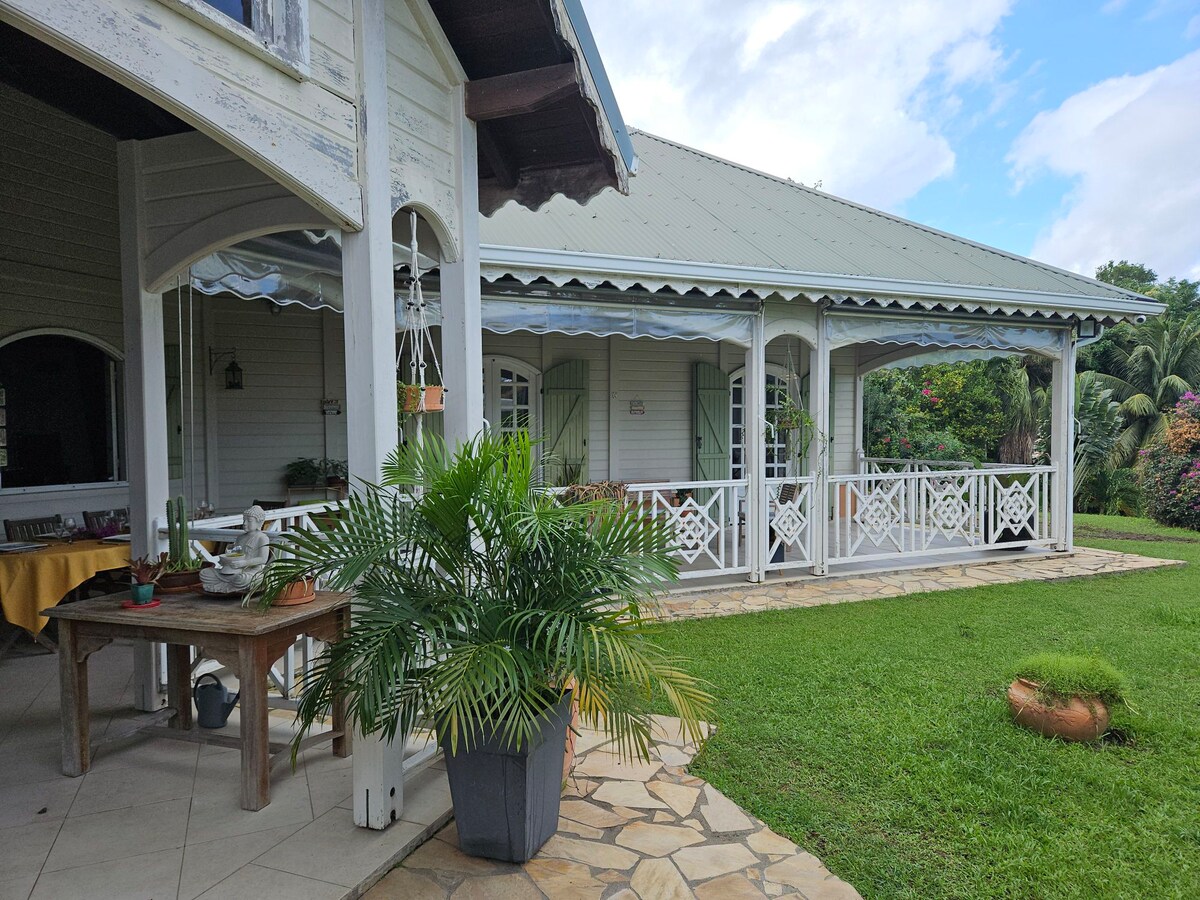 The image depicts a charming colonial-style villa with a covered porch, showcasing decorative railings. A large potted plant adds greenery to the entrance, while a stone pathway leads to a well-maintained lawn, enhancing the inviting outdoor space.