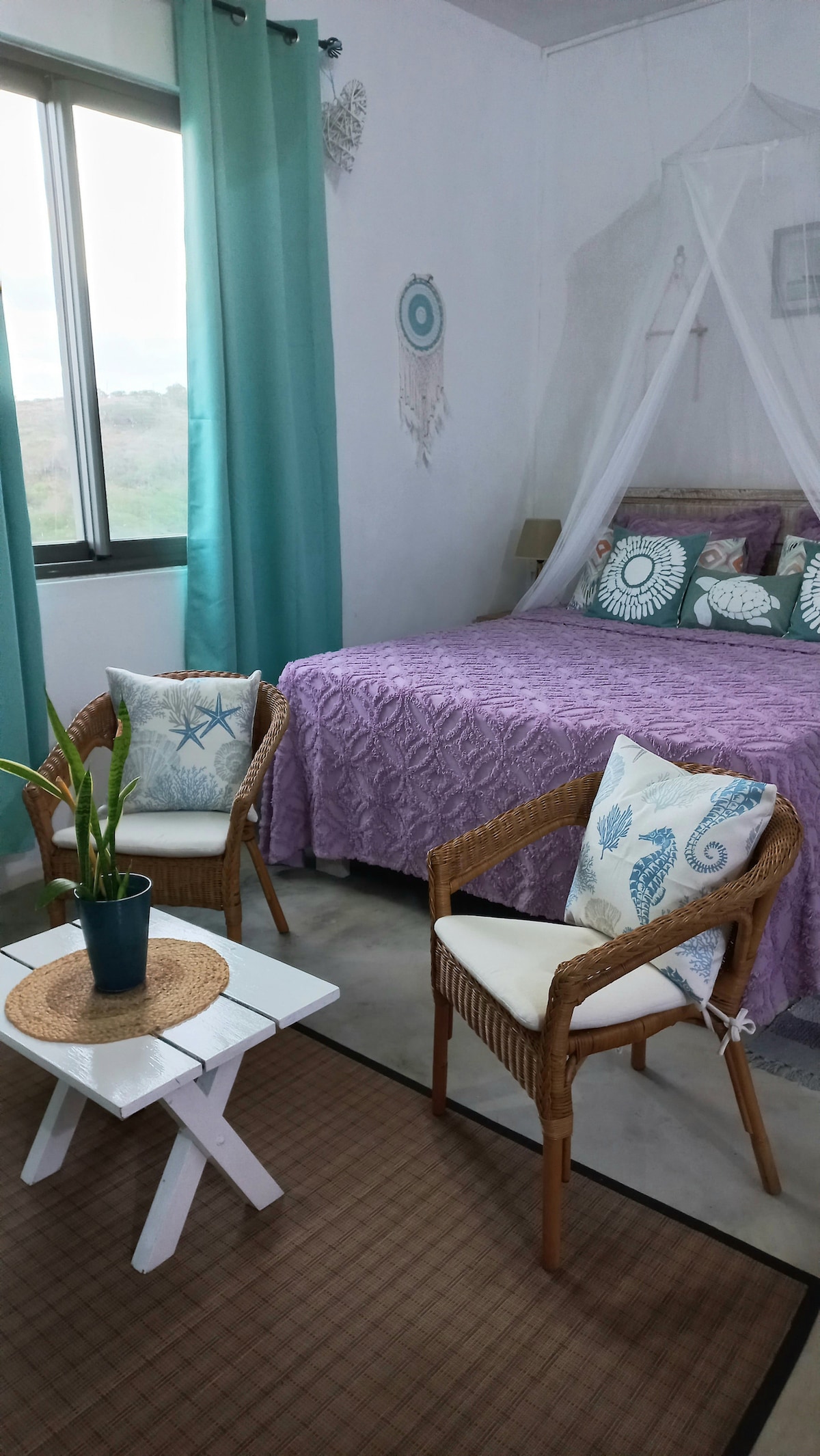 A spacious sleeping area is highlighted by a large bed covered with a purple quilt and decorative pillows featuring marine themes. Two wicker chairs with cushions sit adjacent to a small white table, adding a cozy seating arrangement. Natural light enters through a sizable window framed by teal curtains.