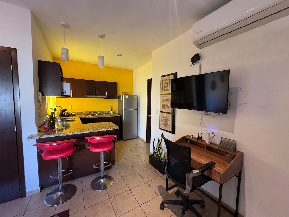 An open kitchen area is shown, featuring modern appliances and a granite countertop. Two red bar stools are positioned at the counter. A work desk with a chair and a mounted television are visible, alongside bright yellow walls that provide a lively contrast.