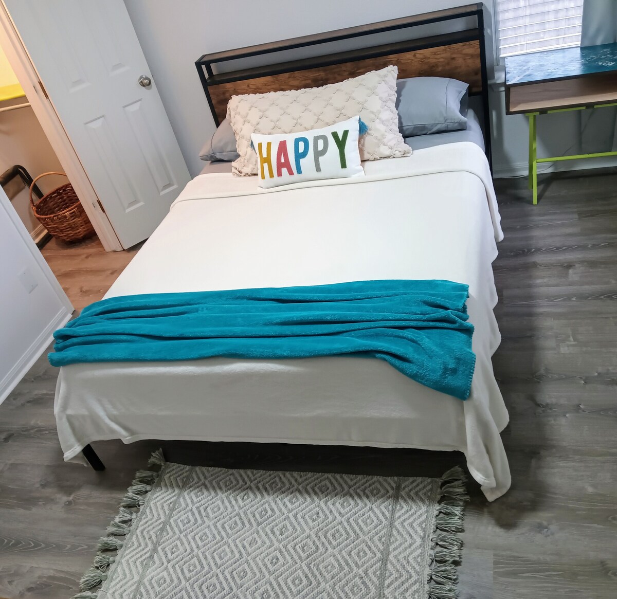 A cozy bedroom features a queen size bed dressed in white linens with a bright blue throw blanket neatly draped across the foot. A decorative pillow with the word 'HAPPY' adds a cheerful touch. A textured area rug lies underneath, complementing the wood-style flooring.