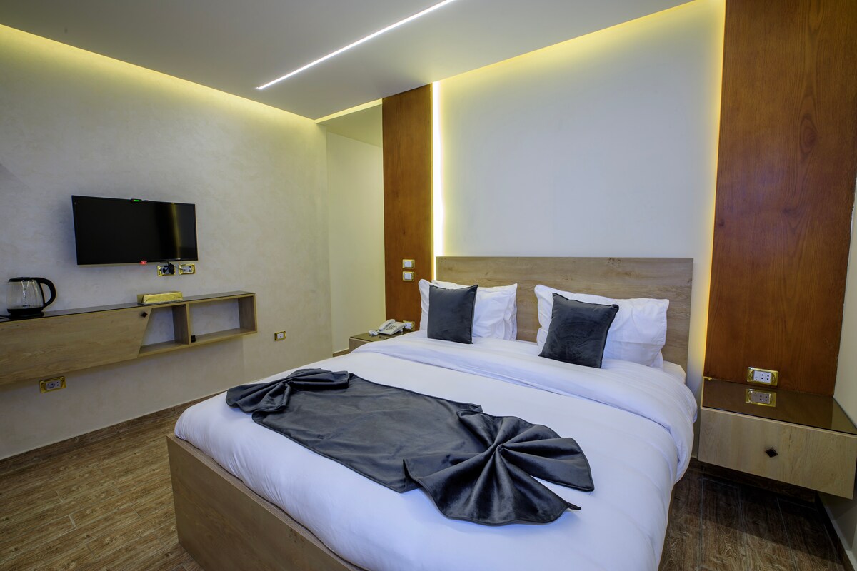 A modern bedroom is presented with soft lighting highlighting a spacious bed adorned with decorative pillows. A flat-screen television is mounted on the wall opposite the bed. A kettle and bedside tables are also visible, contributing to a functional space.