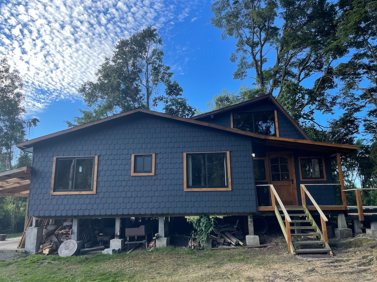 The exterior of a modern cabin is displayed, featuring a blue-grey facade with wooden accents and large windows. A welcoming entrance is visible with a staircase leading up to the front door. Surrounding greenery enhances the natural setting.