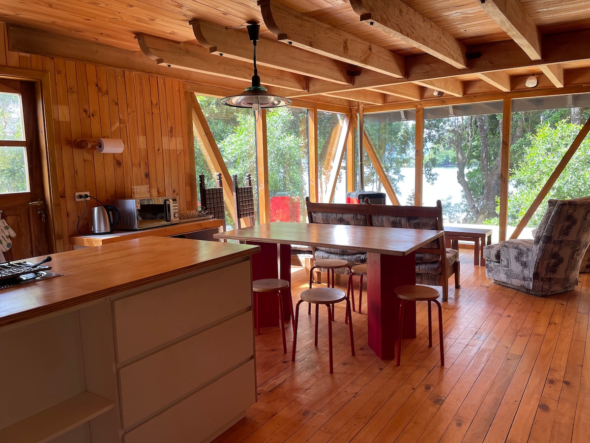 An open living area features wooden beams and a warm wooden floor. A dining table is surrounded by simple chairs, with a cozy seating arrangement nearby. Large windows invite natural light and provide views of the surrounding greenery and the lake beyond.