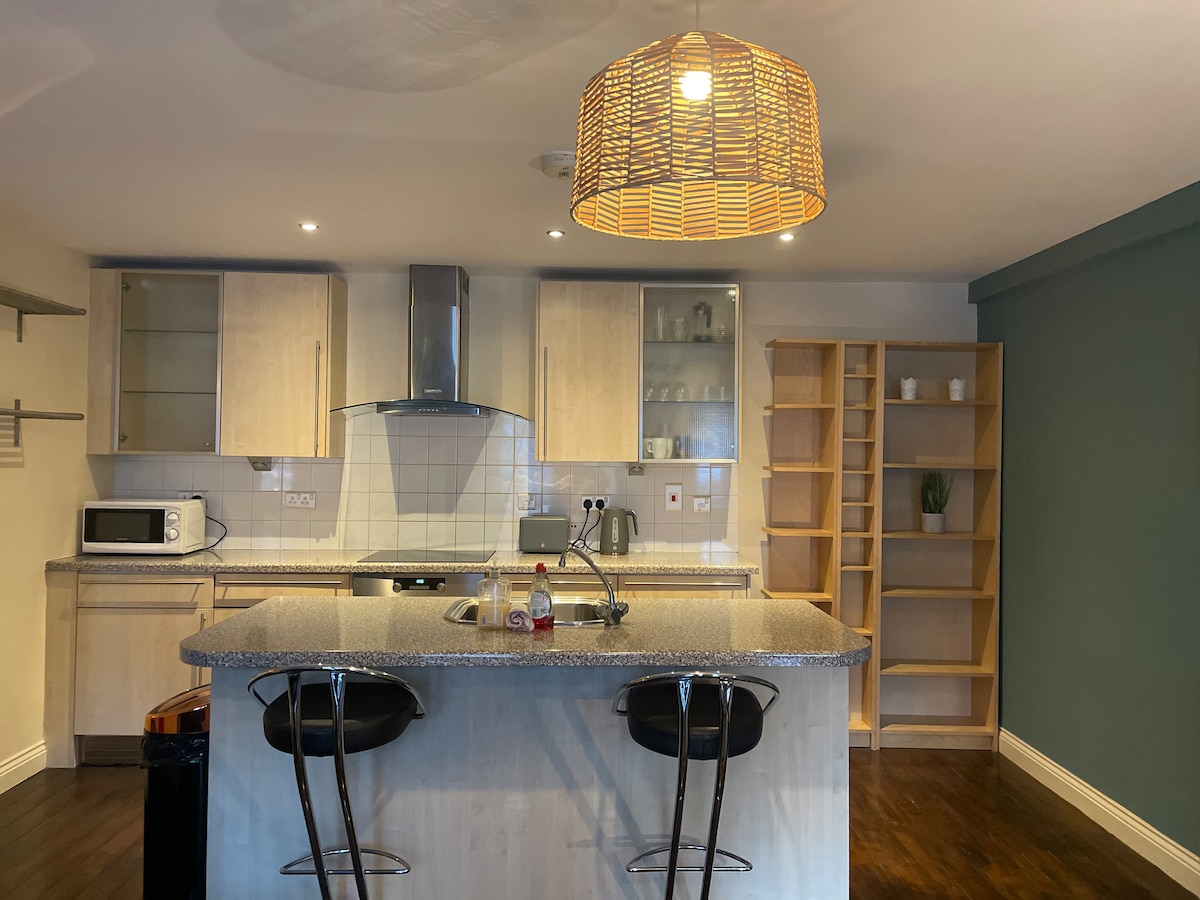 A modern kitchen area features light wooden cabinetry and a spacious island with bar seating. A large woven pendant light hangs from the ceiling, providing illumination. The room includes essential appliances, and there is a wooden shelf unit against the wall, enhancing the space.