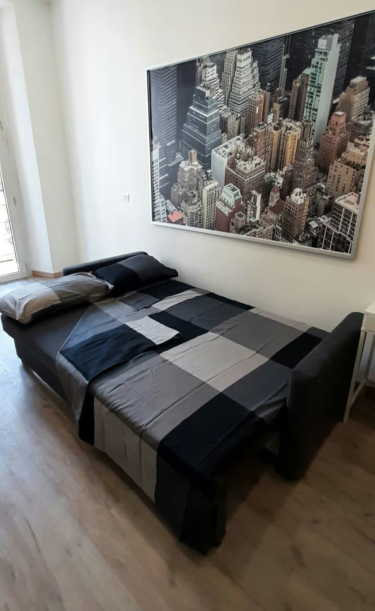 A neatly arranged sleeping area features a spacious bed covered with a patchwork duvet in shades of grey and black. A large wall art piece depicting a cityscape hangs above the bed, while natural light filters through a large window beside it, leading to a balcony.