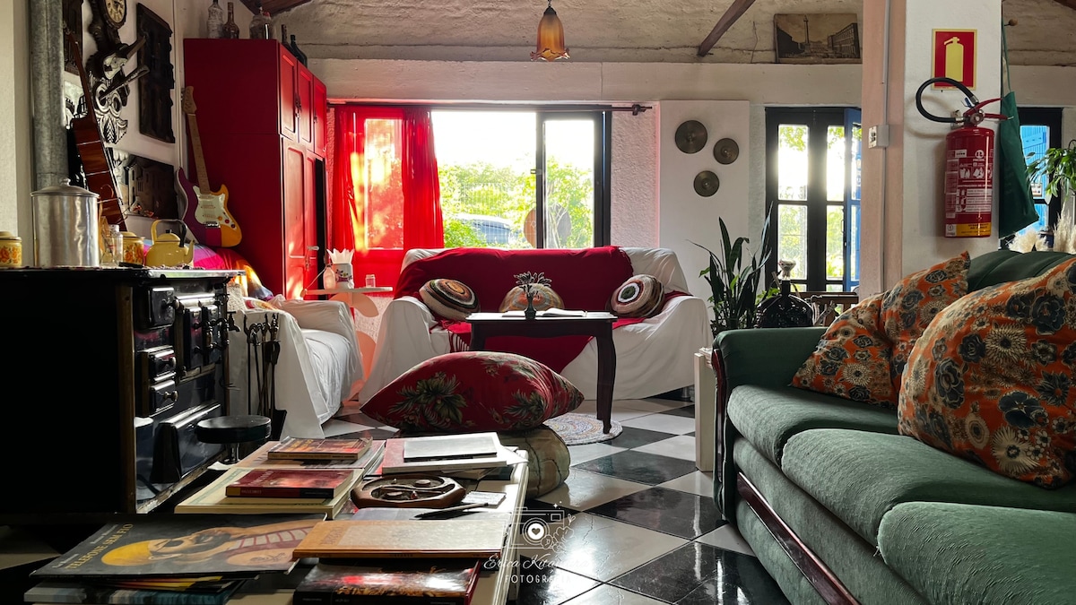 A spacious communal living area features colorful decor and a mix of seating options, including a red sofa, a green couch, and patterned cushions. Large windows invite natural light, while a cozy atmosphere is created by various artworks and plants placed thoughtfully throughout the space.