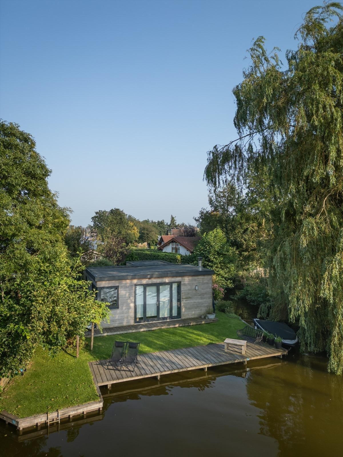 A modern, detached house is set beside a calm waterway, surrounded by lush greenery. Large windows offer views of the serene outdoors, while a small deck extends over the water. The peaceful outdoor area includes chairs for relaxation.