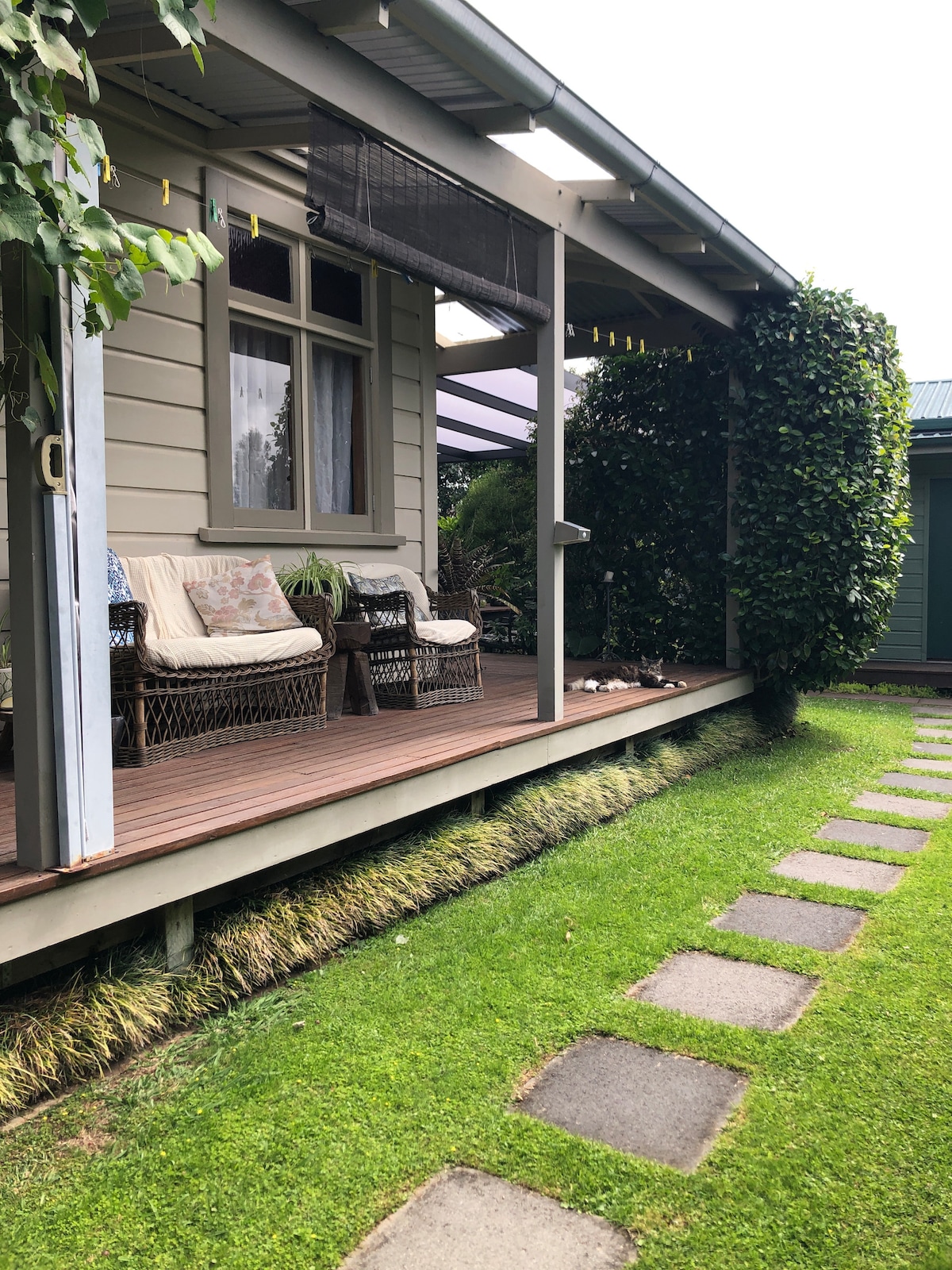 A compact wooden veranda features two wicker chairs with cushions, nestled beneath a shaded overhang. Lush greenery frames the pathway leading to the entrance, with stone stepping stones guiding towards the room. The setting conveys a sense of privacy within the garden landscape.