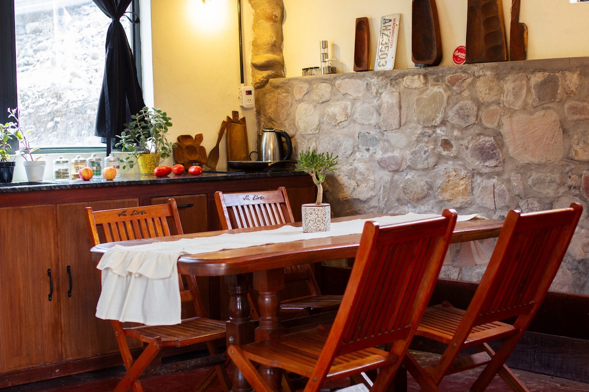 A wooden dining table is surrounded by four stylish folding chairs, inviting communal meals. A stone wall features decorative wooden items, while natural light streams in through a large window adorned with flowing black curtains.