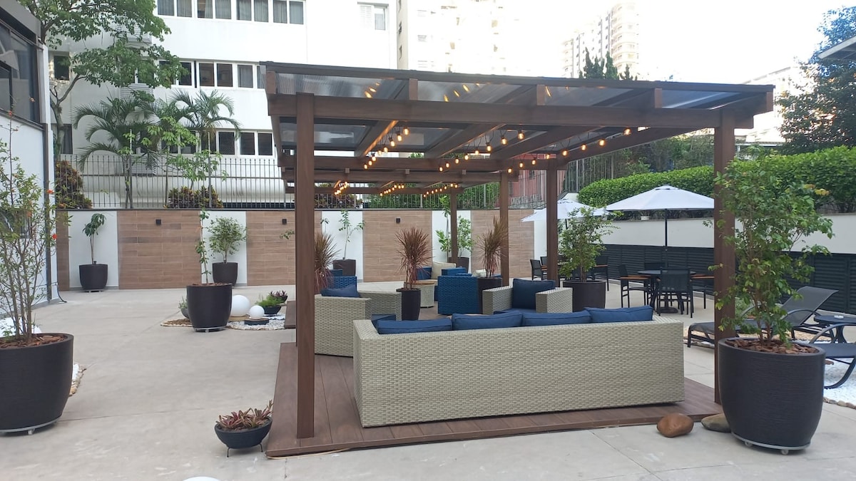 An outdoor lounge area features a wooden pergola with string lights overhead. Comfortable seating is provided by sectional sofas arranged around a central space. Potted plants add greenery, while shaded dining tables are situated in the background, creating a welcoming environment for relaxation.