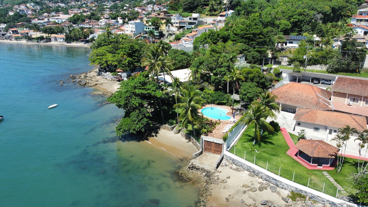 An aerial view showcases a coastal property surrounded by lush greenery and beachfront access. A large swimming pool is positioned near the shore, while sandy beach areas and rocks are visible along the water's edge. The nearby houses and hills create a serene backdrop.