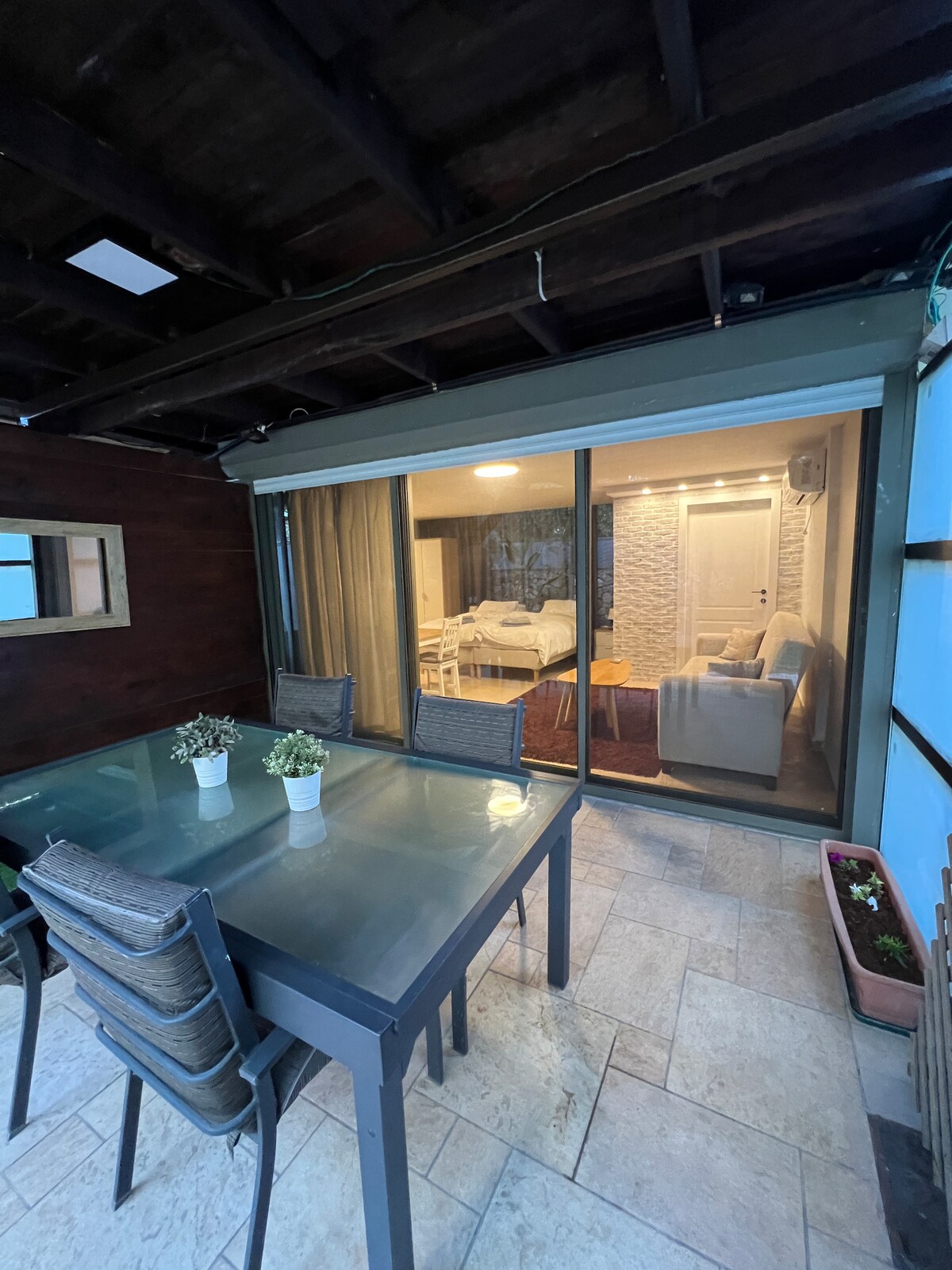 A covered outdoor patio features a glass dining table surrounded by several chairs. Through large sliding glass doors, a cozy living space is visible, showcasing a light-colored sofa and a neatly made bed.