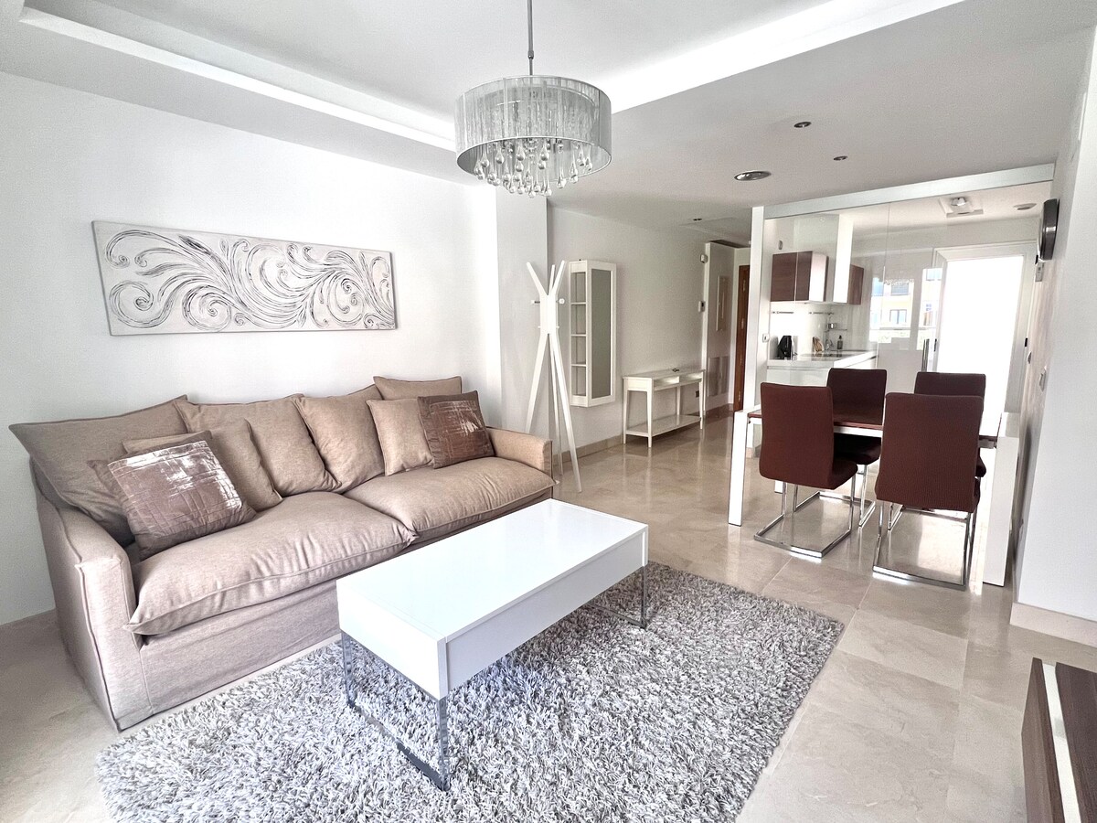 A spacious living area features a large, neutral-colored sofa adorned with decorative cushions, complemented by a modern coffee table. The light-colored walls and tile flooring enhance the room's brightness, while a stylish chandelier and glass dining table add contemporary elegance.